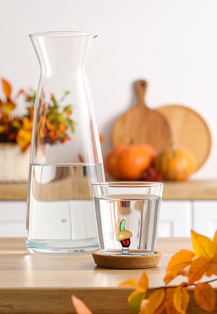 Przezroczysty szklany dzbanek na wodę obok szklanki z korkową podstawą i ozdobną figurką żołędzia, spoczywający na drewnianym stole z jesiennymi liśćmi.