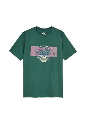 Garçon blond portant un t-shirt vert à motif, un pantalon large foncé et des baskets noires aux semelles blanches, debout les mains dans les poches.