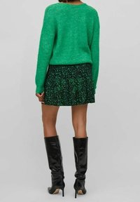 Pull vert duveteux associé à une minijupe noire à motifs floraux et des bottes montantes noires. Texture lisse et silhouette ajustée.