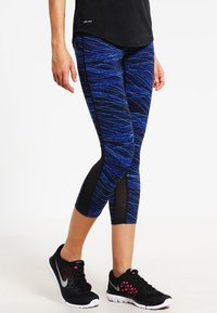 Leggins capri blu e neri con motivo e dettagli in mesh, realizzati in materiale elastico e traspirante; abbinati a scarpe da ginnastica nere.