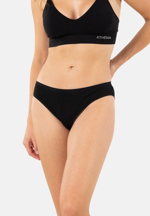 Femme portant un soutien-gorge noir avec le logo « ATHENA » et une culotte assortie noire, posant devant un fond clair uni, montrant le milieu du corps et le haut des cuisses.