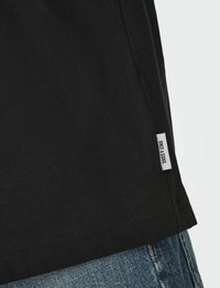 Schwarzes Baumwoll-T-Shirt mit geradem Schnitt, Seitennaht und einem kleinen gewebten Etikett. Die Textur ist glatt, mit sichtbaren Nähdetails.
