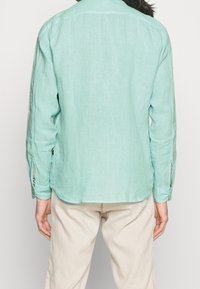 Persona che indossa una camicia in lino verde chiaro a maniche lunghe e pantaloni beige, vista da dietro su uno sfondo bianco uniforme.