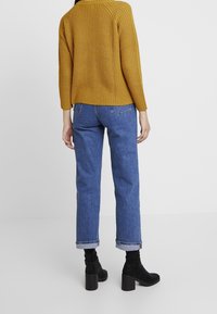 Senffarbenes Rippstrickpullover, lockere Passform; blaue Jeans mit umgekrempelten Bündchen; schwarze Stiefeletten mit Absatz. Einfaches Design ohne sichtbare Muster.
