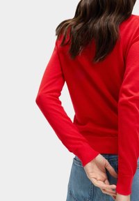 Maglione rosso lavorato a maglia con maniche lunghe, polsini e orlo a costine, caratterizzato da un design semplice. Indossato con jeans a vita alta, presenta un look casual.