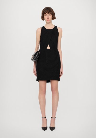 Un vestido negro sin mangas con un corte en forma de llave en la cintura, tejido texturizado y una silueta ajustada. Combinado con un pequeño bolso negro.