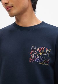 Sweatshirt bleu marine présentant un graphique abstrait coloré sur la poitrine gauche, avec un col côtelé et un tissu doux et texturé.