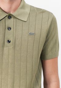 Olivgrön stickad pikétröja med horisontellt randmönster, tre svarta knappar och en liten broderad logotyp på bröstet. Korta ärmar.