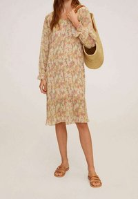 Blumenkleid mit Falten in sanften Farben, langen Ärmeln und Rüschenmuffen. Kombiniert mit einer Strohtasche und braunen Slide-Sandalen.