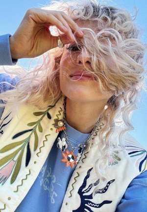 Person with curly light hair partially covering face, wearing a blue shirt, embroidered vest, and colorful sun and flower necklace against blue sky.