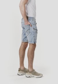 Shorts en jean bleu clair, avec revers roulés, poches avant et arrière, et une texture légèrement délavée. Associés à des chaussures de sport beiges.
