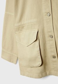 Béžová bavlněná bunda s knoflíkovým zapínáním, dlouhými rukávy a velkou přední kapsou s kulatou klopou.