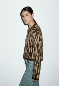 Cardigan a righe marroni e crema in morbido tessuto peloso, con scollatura a V e vestibilità corta, abbinato a jeans azzurri chiari.