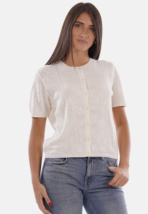 Mujer que lleva una chaqueta de punto blanca de manga corta con botones y un sutil patrón floral, y vaqueros azules de talle alto, de pie con una mano en el bolsillo.
