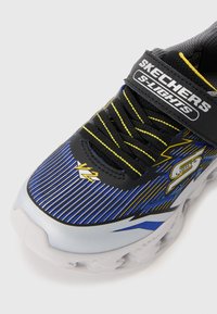Baskets noires et bleues avec un upper en mesh, des accents jaunes, des rayures texturées et une semelle en caoutchouc blanche. Équipées d'une sangle Velcro et de lacets.
