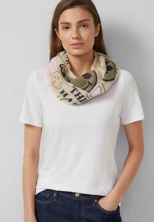 Młoda kobieta w białym t-shircie i kolorowym szaliku z uśmiechniętymi buźkami i tekstem, pozująca z jedną ręką w kieszeni na szarym tle.