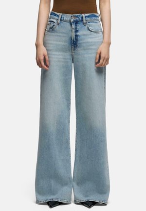 Person iført højtaljede, lyseblå brede jeans med frontlommer, knap- og lynlåslukning, kombineret med spidse sorte sko.