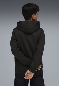 Sudadera negra de tela suave con un bolsillo canguro, puños y dobladillo acanalados; llevada por un niño con las manos entrelazadas detrás.