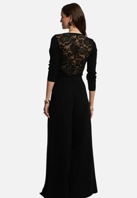 Haut noir à manches longues avec un dos en dentelle transparente et une texture côtelée. Associé à un pantalon noir à taille haute et à jambes larges. Accent en bijoux dorés visibles.