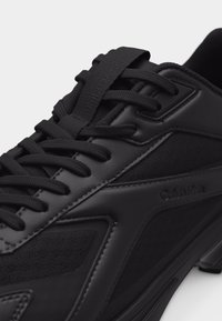 Κοντινό πλάνο μαύρου δερμάτινου και διχτυωτού αθλητικού παπουτσιού με μαύρα κορδόνια και λογότυπο Calvin Klein στο πλάι και στη γλώσσα.