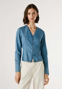 Denim jaka ar V veida kakla, garām piedurknēm un pogu aizdari. Gaiši zila krāsa, ar redzamām šuvēm un nodilušu apakšmalu.