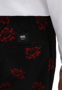 Pantalon noir avec un motif de roses rouges, doté d'une poche arrière, d'une ceinture élastique et d'une étiquette Vans. Le tissu semble doux avec une texture lisse.