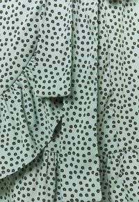Tejido verde con pequeño estampado floral negro. Presenta acentos con volantes y pliegues para textura y detalle de diseño. Material suave.