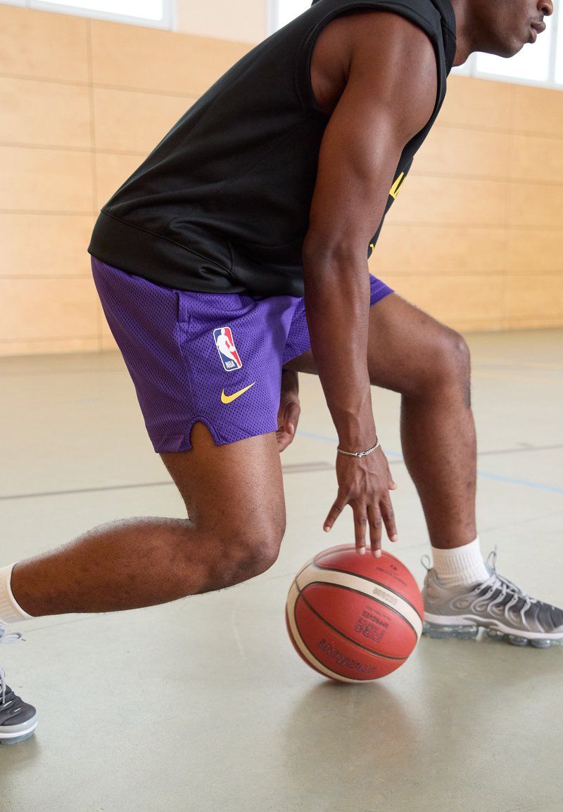 Lilla võrkpallivõrkudega lühikesed püksid Nike ja NBA logodega, must ilma varrukateta särk ja hallid spordijalatsid, palli dribbleerides.