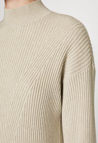 Beige geripptes Rollkragenpullover mit vertikalen Linien, weichem Textur und tailliertem Design. Hoher Kragen und strukturierte Schultern.