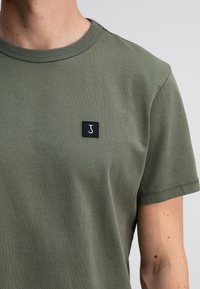 Man in een olijfgroene crew neck t-shirt met een kleine zwarte vierkante patch met daarop een witte letter "J" op de borst.
