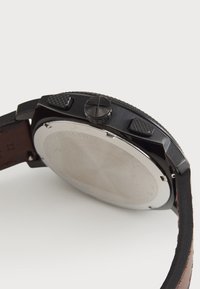 Montre en acier inoxydable noire avec un bracelet en cuir, boutons texturés et boîtier arrière lisse. Elle présente une forme ronde avec des accents contrastés.