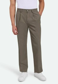 Pantalones de hombre de color verde oliva con un corte relajado, que cuentan con dos bolsillos frontales, un diseño plisado y una textura suave, combinados con zapatillas blancas.