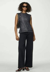 Siva čipkasta bluzica s valovitim rubovima, u kombinaciji s širokim crnim hlačama i crnim sandalama na blok petu. Model drži crnu torbicu.