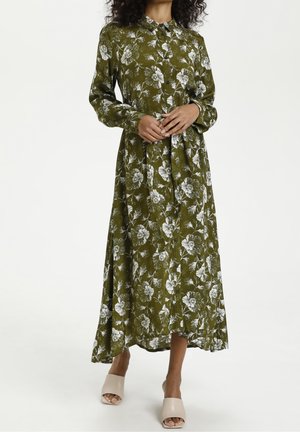 Femme portant une robe midi boutonnée à manches longues à motif floral couleur vert olive avec des sandales beiges à talons blocs et à bouts ouverts, debout sur un fond blanc.