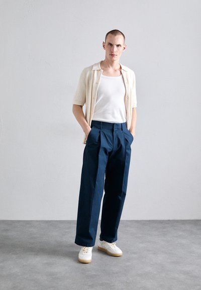 Cardigan en crème à manches courtes et en côtes sur un débardeur blanc, associé à un pantalon taille haute bleu marine et des baskets blanches avec des semelles en gomme.