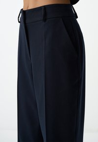 Marineblauwe high-waisted broek met plooien, zijzakken en een zachte textuur. De stof lijkt gestructureerd met een strakke afwerking.
