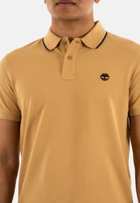 Polo beige en coton texturé, avec un col noir et un petit logo noir sur la poitrine, patte de boutonnage.