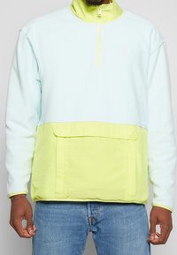 Pullover color blockazzato in blu chiaro e verde neon con mezzo zip, parte superiore in pile e una grande tasca frontale. Texture morbida e vestibilità rilassata.