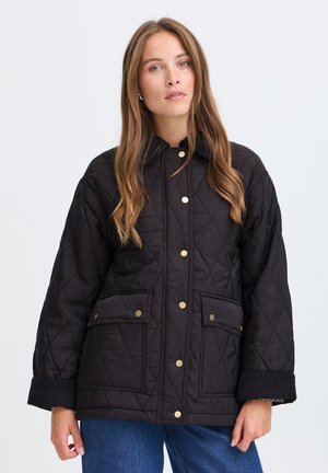 Kvinde med langt hår iført en sort quiltet jakke med guldknapper og store forlommer, står foran en ensfarvet lys baggrund.