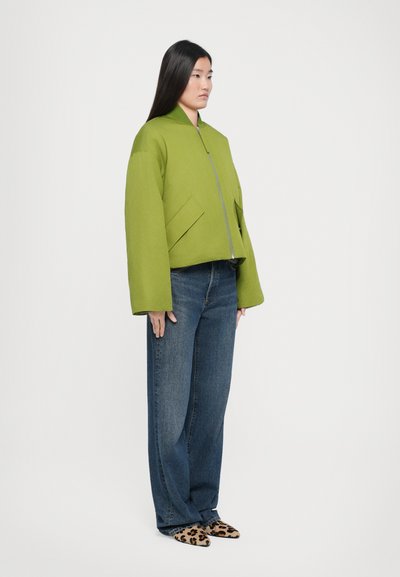 Green cropped jacket with a stand-up collar, front zipper, and two angled pockets. Paired with loose blue jeans and leopard-print shoes.
