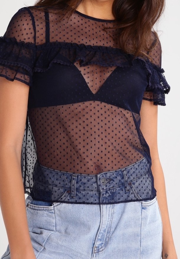 Top in rete trasparente navy con motivo a pois, dettagli arricciati al collo e alle maniche, sovrapposto a un bralette scuro, abbinato a jeans chiari.