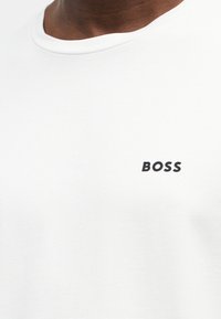 Detailní záběr bílé trička s kulatým výstřihem, na kterém je na levé horní části hrudi černě vytištěné slovo „BOSS“.