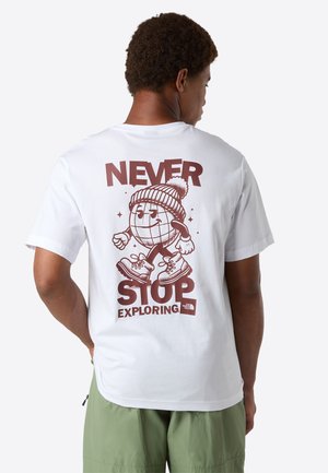 Person iført hvid T-shirt med tegneserieverden, der går i støvler, med teksten "Never Stop Exploring" på ryggen.