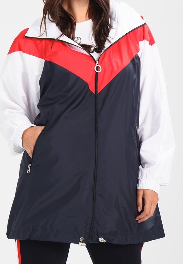 Femme portant un coupe-vent long zippé avec des blocs de couleur rouge, blanc et bleu marine, les mains dans les poches avant.