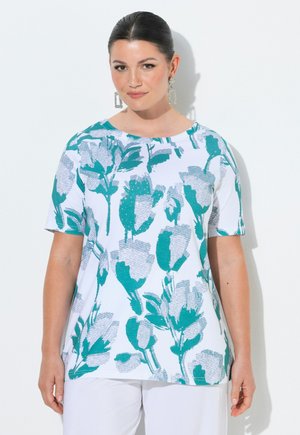 Kvinde med mørkt hår i en lav knold iført en hvid kortærmet skjorte med store turkis- og grå blomsterprint og hvide bukser, står mod en hvid væg.