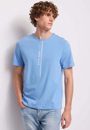 Giovane uomo con capelli ricci che indossa una maglietta azzurra Gaudi Jeans e jeans azzurri, che guarda verso destra con le mani in tasca.