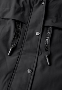 Giacca impermeabile nera con texture liscia, bottoni a pressione e coulisse regolabili con il testo "ESPLORA DI PIÙ".