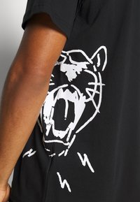 Czarna bawełniana koszulka z dużym nadrukiem białego tygrysa na plecach, z wyrazistymi liniami i dynamicznymi akcentami. Krótkie rękawy i klasyczny krój.