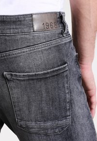 Vista trasera de una persona que lleva puestos unos jeans de mezclilla negra desgastados con un bolsillo trasero cuadrado y un parche de cuero cosido etiquetado como "1968" en la cintura.