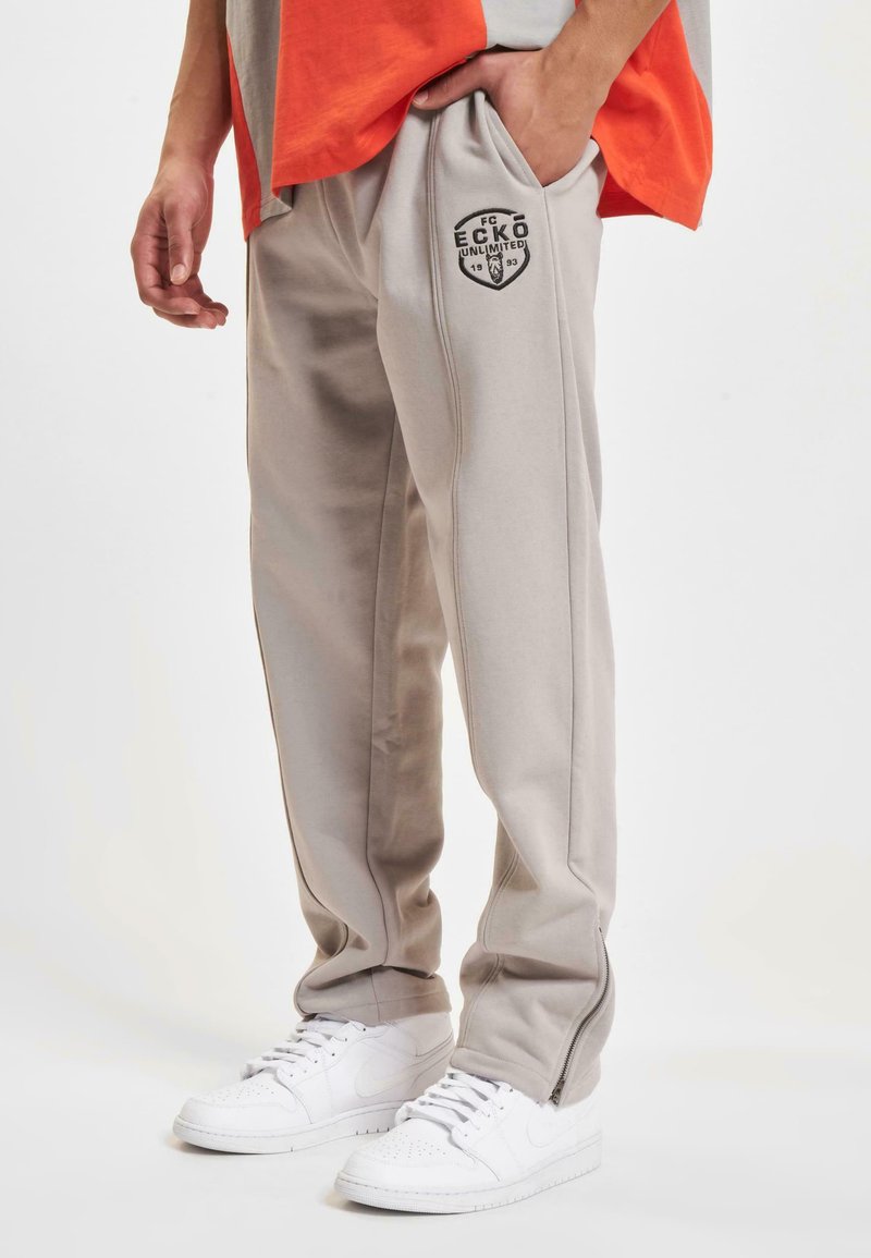 Lys grå joggebukser med glidelås på anklene, sømdetaljer og en svart logo på låret. Kombinert med hvite joggesko.